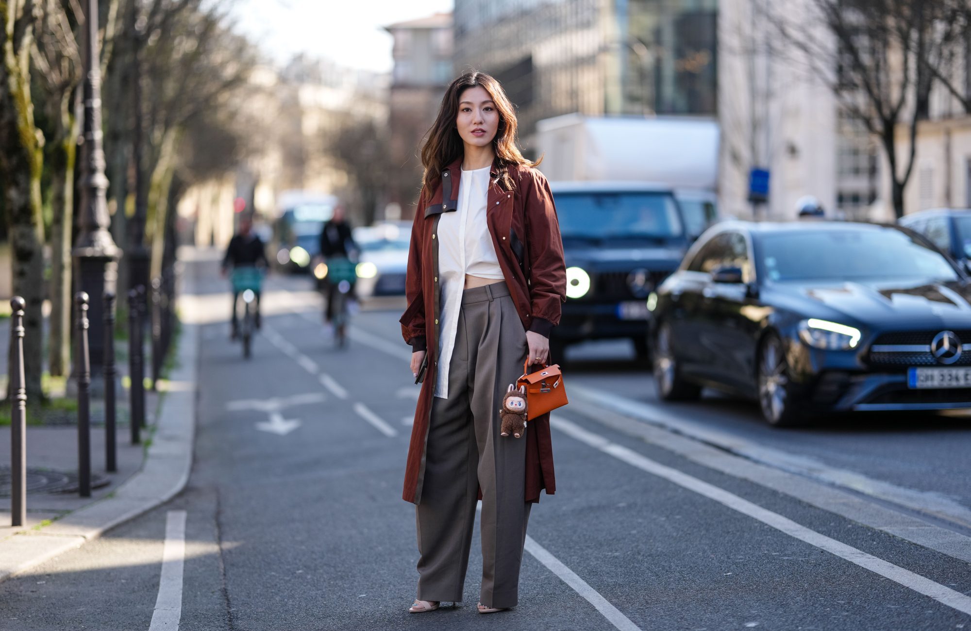 PARIS, FRANCE - MARCH 08: Cristine Sun wears burgundy bordeaux trench coat, white shirt, light gray high waisted trouser pants, shiny dark orange Hermès Kelly leather bag, dark brown Labubu charm, outside Hermès, during the Paris Fashion week Women's Fall/Winter 2025-2026 on March 8, 2025 in Paris, France. (Photo by Edward Berthelot/Getty Images)