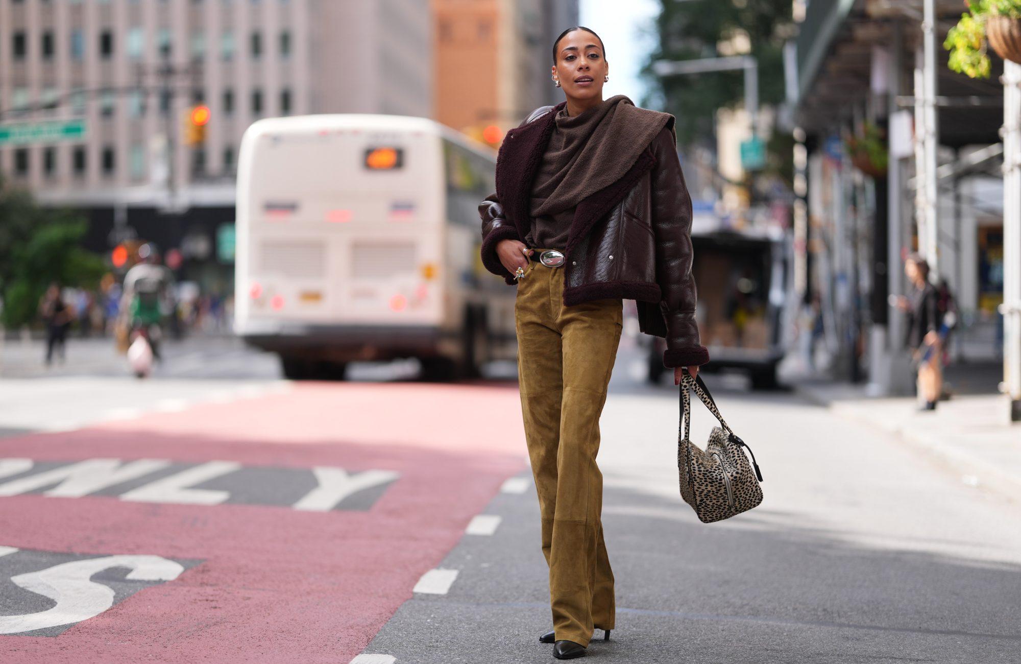 NEW YORK, NEW YORK - SEPTEMBER 12: Olivia Galli wears dark brown slicked back hair in a low bun, a burgundy shearling leather jacket, a brown wool draped cowl-neck top, a suede camel straight-leg pants, a wide tan belt with an oval silver buckle, a beige tote bag with black leopard print and double top handles, black pointed-toe high heels ankle boots shoes, outside Altuzarra, during New York Fashion Week, on September 12, 2026 in New York, New York (Photo by Edward Berthelot/Getty Images)